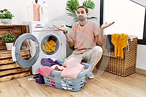 Young hispanic man putting dirty laundry into washing machine clueless and confused expression with arms and hands raised