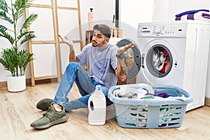 Young hispanic man putting dirty laundry into washing machine clueless and confused expression with arms and hands raised