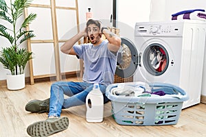 Young hispanic man putting dirty laundry into washing machine crazy and scared with hands on head, afraid and surprised of shock