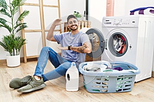 Young hispanic man putting dirty laundry into washing machine gesturing with hands showing big and large size sign, measure symbol