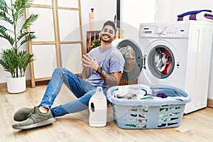 Young hispanic man putting dirty laundry into washing machine hands together and fingers crossed smiling relaxed and cheerful