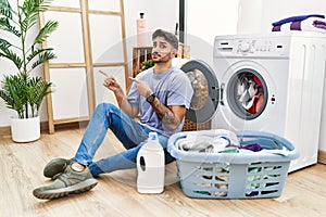 Young hispanic man putting dirty laundry into washing machine pointing aside worried and nervous with both hands, concerned and