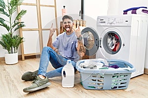 Young hispanic man putting dirty laundry into washing machine shouting with crazy expression doing rock symbol with hands up