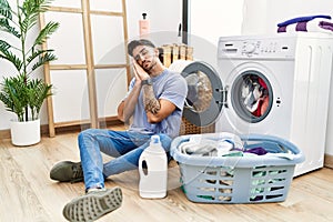 Young hispanic man putting dirty laundry into washing machine sleeping tired dreaming and posing with hands together while smiling