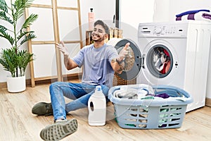 Young hispanic man putting dirty laundry into washing machine smiling cheerful offering hands giving assistance and acceptance