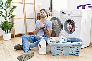 Young hispanic man putting dirty laundry into washing machine smiling with hands on chest with closed eyes and grateful gesture on