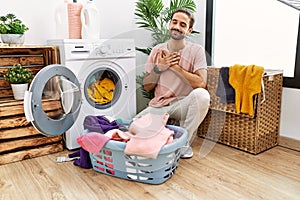 Young hispanic man putting dirty laundry into washing machine smiling with hands on chest with closed eyes and grateful gesture on