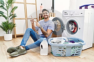 Young hispanic man putting dirty laundry into washing machine smiling and looking at the camera pointing with two hands and