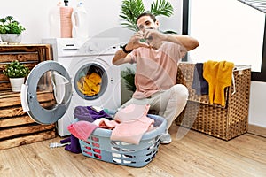 Young hispanic man putting dirty laundry into washing machine smiling in love showing heart symbol and shape with hands