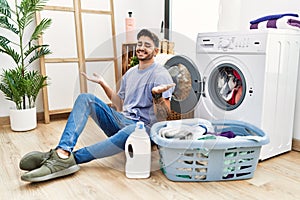 Young hispanic man putting dirty laundry into washing machine smiling showing both hands open palms, presenting and advertising