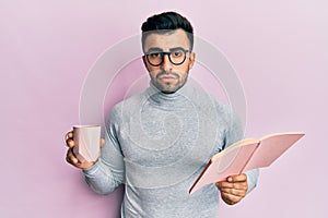 Young hispanic man reading a book and drinking a cup of coffee depressed and worry for distress, crying angry and afraid