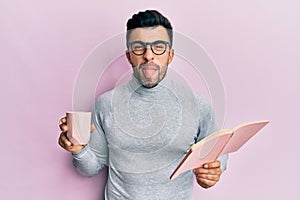 Young hispanic man reading a book and drinking a cup of coffee sticking tongue out happy with funny expression