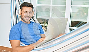 Young hispanic man relaxed working using laptop and headphones lying on the hammock at terrace