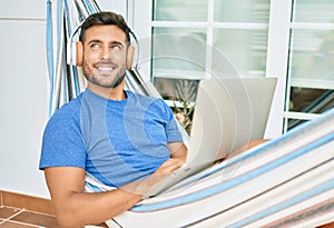 Young hispanic man relaxed working using laptop and headphones lying on the hammock at terrace