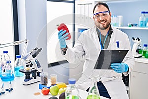 Young hispanic man scientist write on report working holding red pepper at laboratory