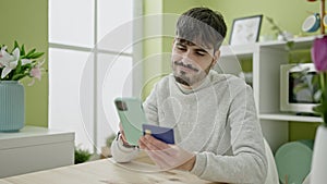 Young hispanic man shopping online with smartphone sitting on the table at dinning room