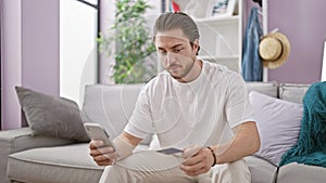 Young hispanic man shopping with smartphone and credit card looking upset at home