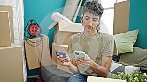 Young hispanic man shopping with smartphone and credit card sitting on sofa at new home