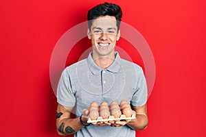 Young hispanic man showing fresh white eggs looking positive and happy standing and smiling with a confident smile showing teeth