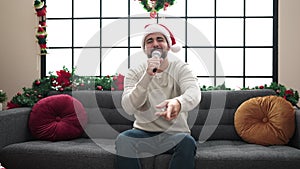 Young hispanic man singing sitting on sofa by christmas decoration at home