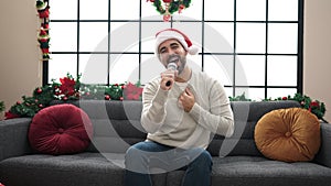 Young hispanic man singing sitting on sofa by christmas decoration at home