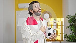 Young hispanic man singing soccer team anthem with hand on heart holding football ball at home