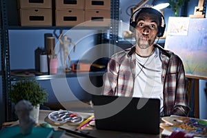 Young hispanic man sitting at art studio with laptop late at night making fish face with lips, crazy and comical gesture