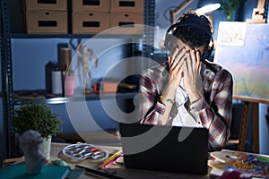 Young hispanic man sitting at art studio with laptop late at night with sad expression covering face with hands while crying