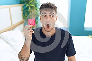 Young hispanic man sitting on the bed holding condom scared and amazed with open mouth for surprise, disbelief face