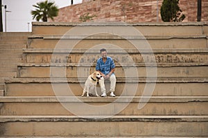 Young Hispanic man, sitting on big cement stairs next to his dog laughing very happy. Concept, dogs, pets, animals, friends