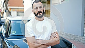 Young hispanic man sitting on car with arms crossed gesture and serious face at street