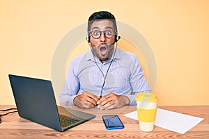 Young hispanic man sitting at the desk wearing operator headset at the call center office scared and amazed with open mouth for