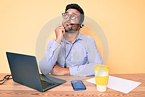 Young hispanic man sitting at the desk wearing operator headset at the call center office serious face thinking about question