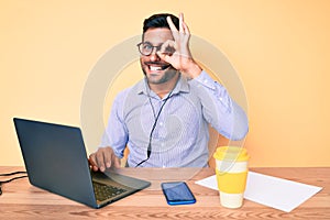 Young hispanic man sitting at the desk wearing operator headset at the call center office smiling happy doing ok sign with hand on