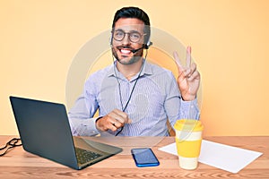 Young hispanic man sitting at the desk wearing operator headset at the call center office smiling with happy face winking at the