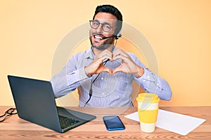 Young hispanic man sitting at the desk wearing operator headset at the call center office smiling in love showing heart symbol and