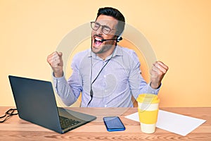 Young hispanic man sitting at the desk wearing operator headset at the call center office very happy and excited doing winner