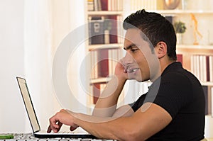 Young hispanic man sitting at desk working on white laptop, profile angle