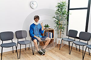 Young hispanic man sitting at doctor waiting room with neck injury smiling happy and positive, thumb up doing excellent and