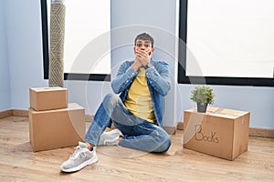 Young hispanic man sitting on the floor at new home shocked covering mouth with hands for mistake