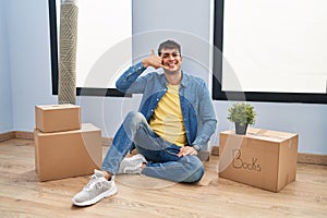 Young hispanic man sitting on the floor at new home smiling doing phone gesture with hand and fingers like talking on the