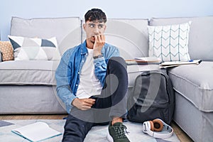 Young hispanic man sitting on the floor studying for university looking stressed and nervous with hands on mouth biting nails