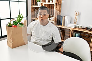 Young hispanic man sitting with paper bag with groceries scared and amazed with open mouth for surprise, disbelief face