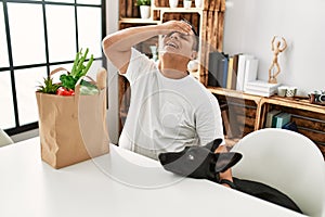 Young hispanic man sitting with paper bag with groceries stressed and frustrated with hand on head, surprised and angry face