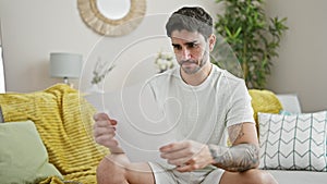 Young hispanic man sitting on sofa breaking paper at home