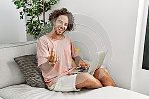 Young hispanic man sitting on the sofa at home using laptop beckoning come here gesture with hand inviting welcoming happy and