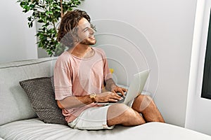 Young hispanic man sitting on the sofa at home using laptop looking away to side with smile on face, natural expression