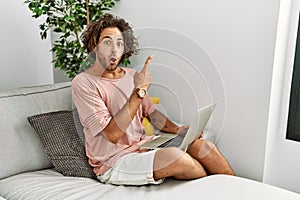 Young hispanic man sitting on the sofa at home using laptop surprised pointing with finger to the side, open mouth amazed