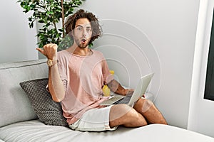 Young hispanic man sitting on the sofa at home using laptop surprised pointing with hand finger to the side, open mouth amazed