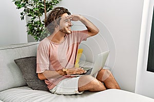 Young hispanic man sitting on the sofa at home using laptop very happy and smiling looking far away with hand over head